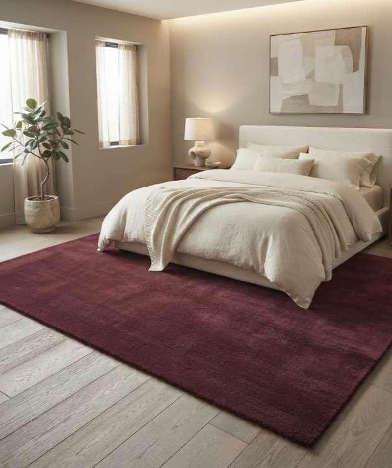 Burgundy wine red area rug in a cozy bedroom setting with a cream upholstered bed, knit throw blanket, walnut nightstands, and soft ambient lighting, highlighting the rug's soft plush pile and warm deep red tone underfoot.