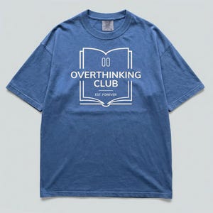 Puede incluir: Una camiseta azul de manga corta con un gráfico blanco de un libro abierto con el texto OVERTHINKING CLUB y EST. FOREVER impreso en el centro. La camiseta tiene cuello redondo y un corte holgado con una etiqueta visible en el cuello.