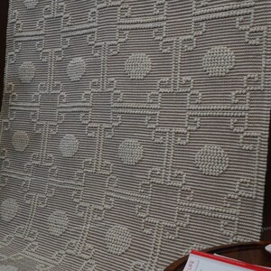 Op de afbeelding: Een beige geweven textiel met een verhoogd, getextureerd patroon van witte cirkelvormige stippen en onderling verbonden geometrische lijnen. De stof vertoont een herhalend rasterontwerp met een consistente, tactiele oppervlaktestructuur.