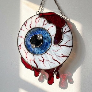 Könnte beinhalten: Ein kreisförmiger Buntglas-Sonnenfänger in Form eines menschlichen Auges mit weißer Sklera, blauer Iris und schwarzer Pupille. Das Stück zeigt rote Glasadern und dunkelrote Glastropfen, die an einer Metallkette aufgehängt sind.