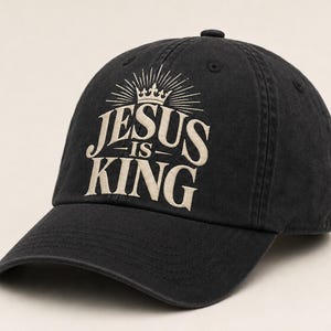 Könnte beinhalten: Eine schwarze Baseballkappe mit einem gestickten Design in hellem Beige. Das Design zeigt eine Krone mit strahlenförmigen Linien über dem Text "JESUS IS KING" in einer fetten Serifenschrift, die auf der Vorderseite der Kappe zentriert ist.