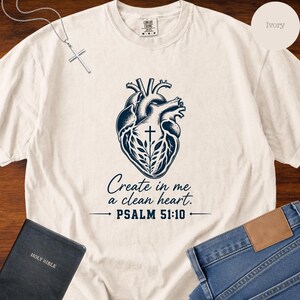Könnte beinhalten: Ein elfenbeinfarbenes Kurzarm-T-Shirt mit einer dunkelblauen Grafik eines anatomischen Herzens, das mit einem Kreuz und Blättern verziert ist. Darunter steht der Text "Create in me a clean heart. PSALM 51:10" in dunkelblauer Schrift.