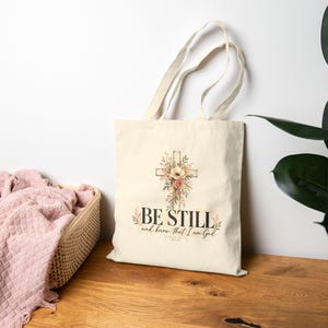 Könnte beinhalten: Eine cremefarbene Stofftasche mit einem Aufdruck, der ein mit Aquarellblumen verziertes Kreuz zeigt. Der Text "BE STILL and know that I am God" ist in schwarzer Schrift gedruckt, darunter steht "Psalm 46:10" in kleinerer Schriftgröße.
