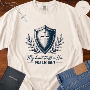 May include: An ivory short-sleeve t-shirt featuring a dark blue graphic of a shield with a cross in the center, flanked by olive branches. Below the shield, the text reads: My heart trusts in Him. PSALM 28:7. The shirt has a crew neckline.