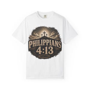 Könnte beinhalten: Ein weißes Kurzarm-T-Shirt mit einer kreisförmigen Grafik auf der Brust. Das Design zeigt drei Kreuze auf einem Hügel unter einem strahlenden Himmel, mit dem Text Philippians 4:13 in beiger Schrift und einer Dornenkrone am unteren Rand.