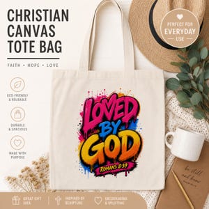 Könnte beinhalten: Eine beigefarbene Canvas-Tragetasche mit einem farbenfrohen Grafikdruck, der den Text "LOVED BY GOD" in einem kräftigen, tropfenden Graffiti-Stil zeigt, sowie der Bibelstellenangabe "ROMANS 8:39" in kleineren schwarzen Buchstaben unter dem Motiv.