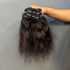 Könnte beinhalten: Ein Bündel dunkelbrauner Haarverlängerungen mit welliger Textur. Das Haar ist am oberen Ende mit einem dicken, vernähten Tressenband befestigt, das durch mehrere kleine weiße Kunststoffbänder zusammengehalten wird, um einen festen Halt zu gewährleisten.