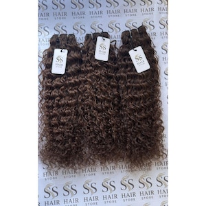 Puede incluir: Tres paquetes de extensiones de cabello rizado de color marrón oscuro, cada uno con una etiqueta rectangular blanca con el texto SS HAIR STORE impreso en negro. El cabello tiene un patrón de rizos apretado y uniforme dispuesto verticalmente.