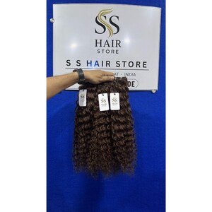 K&ouml;nnte beinhalten: Ein B&uuml;ndel dunkelbrauner, lockiger Haarverl&auml;ngerungen mit drei wei&szlig;en, rechteckigen Etiketten, die jeweils das SS HAIR STORE-Logo tragen. Das Haar wird vor einem wei&szlig;en Schild mit dem Text SS HAIR STORE und SURAT - INDIA gehalten.