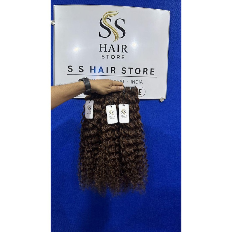 K&ouml;nnte beinhalten: Drei B&uuml;ndel dunkelbrauner, lockiger Haarverl&auml;ngerungen mit einer engen, gewellten Textur. Jedes B&uuml;ndel hat ein wei&szlig;es, rechteckiges Etikett mit dem Text SS HAIR STORE. Das Haar wird vor einem wei&szlig;en Schild mit dem Logo SS HAIR STORE gezeigt.