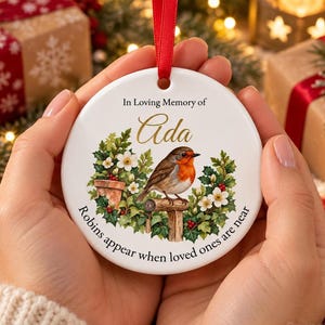 May include: A round white ceramic ornament with a red ribbon hanger. The design features a robin perched on a wooden post surrounded by green leaves and white flowers. Text reads: In Loving Memory of Ada, Robins appear when loved ones are near.