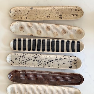 May include: Six elongated ceramic trays arranged in a vertical stack. The trays feature various surface textures and patterns, including brown speckles, vertical ridges, black stripes, and a dark brown glaze, all with rounded ends and edges.