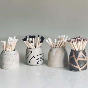 May include: Four small ceramic vessels holding wooden matches. The first is speckled grey, the second is cream with black squiggles, the third is a smooth beige with a white drip glaze, and the fourth is dark grey with a tan geometric pattern.