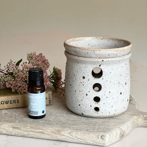 May include: A speckled white ceramic oil warmer with a cylindrical body and four vertical circular cutouts, accompanied by a small amber glass bottle labelled Rebecca Tracey Shanti botanical oil blend with a black cap.