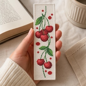 Può includere: Un segnalibro rettangolare con un motivo a punto croce raffigurante ciliegie rosse con foglie verdi su uno sfondo di tessuto bianco. Il design include diversi grappoli di ciliegie collegati da sottili steli verdi e piccoli punti rossi.