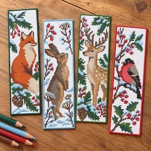 May include: Four rectangular cross-stitch bookmarks featuring winter nature scenes. One shows a fox, one a hare, one a deer, and one a bullfinch bird. Each bookmark is decorated with pine branches, holly berries, and snow, finished with coloured borders.