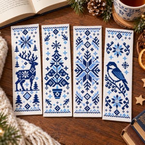 May include: Four rectangular bookmarks featuring blue and white cross-stitch patterns. The designs include a deer, a potted Christmas tree, a large snowflake, and a bird perched on a branch, all set against a light-coloured fabric background.