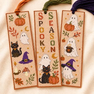 May include: Three rectangular cross-stitch bookmarks on beige fabric with orange, green, and dark purple tassels. Designs include ghosts, black cats, pumpkins, witch hats, mushrooms, and leaves. The center bookmark features the text SPOOKY SEASON.