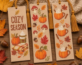May include: Three rectangular fabric bookmarks with cross-stitch embroidery featuring autumn themes. The designs include pumpkins, mushrooms, acorns, leaves, and mugs of hot drinks. Each bookmark has a mustard yellow tassel attached to the top.