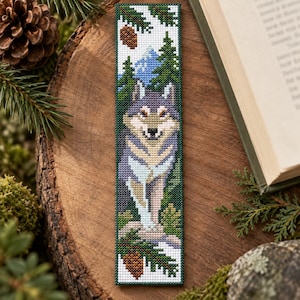 May include: A rectangular bookmark featuring a cross-stitch design of a grey and white wolf standing in a forest. The scene includes green pine trees, blue mountains, and brown pine cones against a white background with a dark green border.