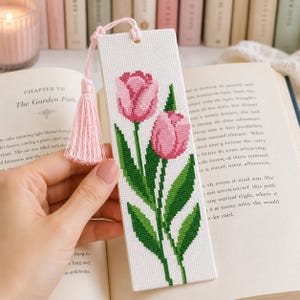 Pode incluir: Um marcador de página retangular branco com um padrão de ponto cruz de duas tulipas cor-de-rosa com caules e folhas verdes. O marcador é feito de tecido e inclui uma borla de fio rosa claro presa no topo através de um pequeno orifício.