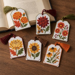 May include: Six rectangular cross-stitch bookmarks featuring autumn-themed floral designs including sunflowers, marigolds, and dahlias. Each white fabric bookmark is finished with a colored embroidery thread tassel attached to a small hole at the top.