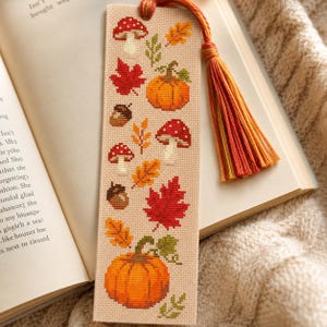 May include: A rectangular bookmark made of beige Aida fabric featuring cross-stitch patterns of red and white mushrooms, orange pumpkins, brown acorns, and red and yellow autumn leaves. A multi-coloured orange, red, and yellow tassel is attached.