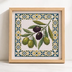 Pode incluir: Um bordado em ponto cruz quadrado apresentando um ramo central com duas azeitonas roxo-escuras e duas azeitonas verde-claras rodeadas por folhas verdes. O design é emoldurado por uma borda geométrica azul, amarela e verde numa moldura de madeira clara.