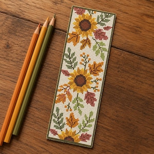 May include: A rectangular cross-stitch bookmark featuring a pattern of yellow sunflowers, orange and red autumn leaves, and green foliage on a white fabric background, accompanied by three colored pencils in orange, yellow, and green.
