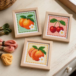 May include: Three square wooden frames containing cross-stitch art of fruit. One features an orange peach on a light green background, another shows two red cherries on a peach background, and the third displays a peach on a yellow background.