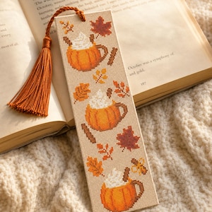 May include: A rectangular cross-stitch bookmark on light beige fabric featuring three orange pumpkin-shaped mugs filled with white whipped cream, surrounded by brown cinnamon sticks and various orange, red, and brown autumn leaves with a rust-colored tassel.