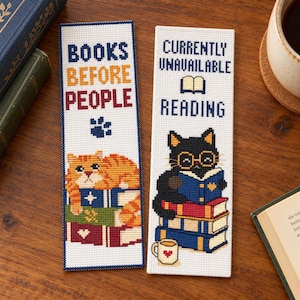 May include: Two rectangular cross-stitch bookmarks on a white fabric base. The left bookmark reads BOOKS BEFORE PEOPLE with an orange tabby cat on books. The right bookmark reads CURRENTLY UNAVAILABLE READING with a black cat wearing glasses.
