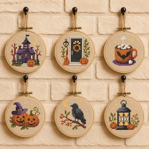May include: Six circular wooden embroidery hoops containing cross-stitch designs featuring Halloween themes including a purple haunted house, a black door, a mug with a bat, two jack-o'-lanterns, a black crow on a branch, and a lantern.