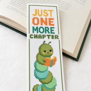 May include: A rectangular white fabric bookmark featuring cross-stitch embroidery. The text reads JUST ONE MORE CHAPTER in yellow, orange, and green. Below the text is a green caterpillar with pink cheeks holding an open orange book.
