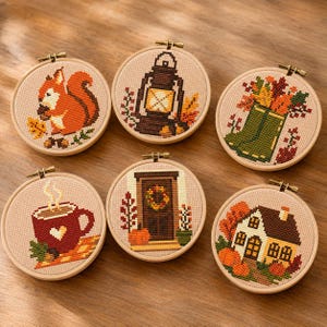 May include: Six circular embroidery hoops featuring cross-stitch designs on beige fabric. The patterns include an orange squirrel, a brown lantern, a green boot with autumn leaves, a red mug with a heart, a brown door with a wreath, and a house.