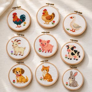 Könnte beinhalten: Neun runde Stickrahmen aus Holz mit Kreuzstichmotiven von Bauernhoftieren, darunter ein Hahn, eine Henne, eine Ente, eine Ziege, ein Schwein, eine Kuh, ein Hund, eine Katze und ein Kaninchen auf weißem Stoff mit kleinen Blumen.