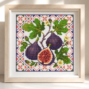 May include: A square cross-stitch embroidery in a light wood frame featuring three figs, one sliced open to reveal a red seeded interior, surrounded by green leaves and small white flowers, all set against a white grid with a red and blue geometric border.