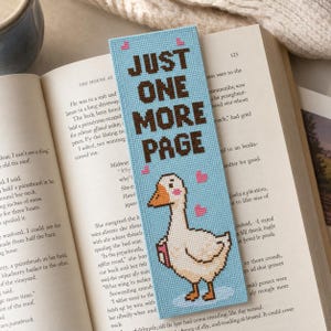 May include: A light blue cross-stitch bookmark featuring the brown text "JUST ONE MORE PAGE" and a white goose with an orange beak and feet, accented by small pink hearts, resting on an open book with printed text on the pages.