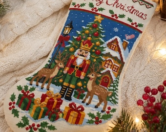 May include: A cream-coloured Christmas stocking featuring a cross-stitched design of a nutcracker, two deer, a decorated Christmas tree, a house, and wrapped gifts. The text Merry Christmas is embroidered at the top with red and green holly accents.