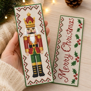 May include: Two rectangular cross-stitch bookmarks on cream fabric. One features a nutcracker in a red, gold, and green uniform with a crown. The other displays the red text Merry Christmas with green holly leaves and red berries, both with red and green borders.