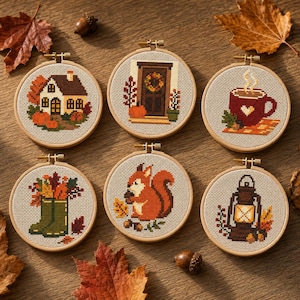 May include: Six circular embroidery hoops featuring cross-stitch designs on light fabric. The patterns include a house, a front door with a wreath, a mug of hot drink, green rain boots with leaves, a squirrel with an acorn, and a lantern.