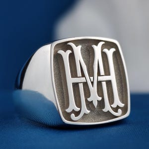 May include: A silver signet ring featuring a square face with rounded corners. The centre of the ring displays a raised, polished monogram of the letters H and M intertwined in a decorative script font against a recessed, matte-textured background.