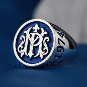 Könnte beinhalten: Ein silberfarbener Siegelring mit einer kreisförmigen Platte und einer tiefblauen Einlage. In der Mitte befindet sich ein filigranes silbernes Monogramm der Buchstaben MP mit einer Lilienverzierung. Die Seite des Rings trägt die Gravur 1974.