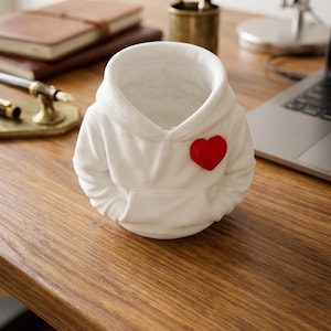 Peut inclure: Un organisateur de bureau en céramique blanche en forme de sweat-shirt à capuche miniature. Le récipient présente une capuche sculptée, des manches longues, une poche kangourou frontale et un petit cœur rouge en relief sur la poitrine gauche.