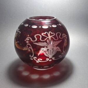 Può includere: Un vaso sferico in vetro rosso rubino caratterizzato da intricati disegni incisi in bianco raffiguranti un cesto pieno di fiori e fogliame, circondato da volute decorative e un motivo ovale ripetuto sui bordi superiore e inferiore.