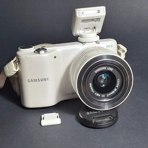Op de afbeelding: Een witte Samsung NX digitale camera met een bevestigde lens, een uitklapbare flitser en een camerariem. De lens is gelabeld met Samsung Lens 1:3.5-5.8 20-50mm II ED Ø40.5 i-Function. Een losse zwarte lensdop en een klein wit kapje zijn inbegrepen.