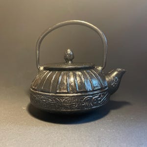 Op de afbeelding: Een zwarte gietijzeren theepot met een afgerond lichaam, verticale ribbels aan de bovenzijde en een ingewikkeld reliëfpatroon van krullen rond de basis. De theepot heeft een bijpassend deksel met een knop en een zwenkbare handgreep.