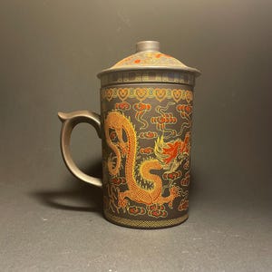 Può includere: Una tazza in ceramica marrone scuro con coperchio abbinato e manico curvo. L'esterno presenta un dettagliato motivo a drago dorato e rosso circondato da nuvole stilizzate e motivi geometrici che bordano i margini superiore e inferiore.