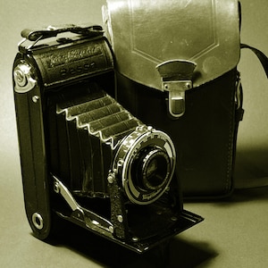May include: A vintage Voigtländer Bessa folding camera with a black bellows and a metal lens assembly, accompanied by a matching brown leather carrying case with a metal clasp. The camera body features the brand name and model embossed on the top.