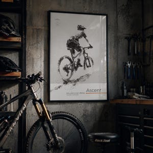 May include: A framed vertical poster featuring a black and white ink-style illustration of a mountain biker on a trail. The text reads Ascent, Every ascent is an offering, and includes small coordinate details. The poster is displayed on a concrete wall.